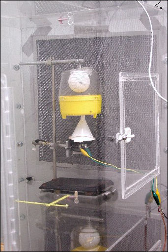 The plexiglass cage is shown, containing a large trap suspended from a metal stand. A funnel, in contact with a small loudspeaker, contacts the trap from below.